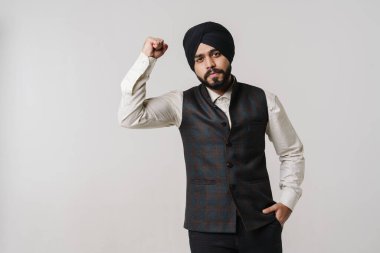 Sarık takan sakallı Güney Asyalı adam, pazılarını beyaz duvarın üzerinde soyutlanmış olarak gösteriyor.