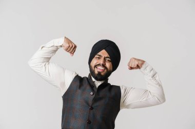 Sarık takan sakallı Güney Asyalı adam kaslarını beyaz duvarın üzerinde soyutlanmış olarak gösteriyor.