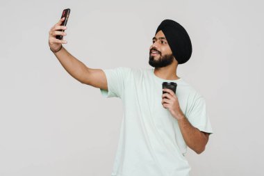 Güney Asyalı adam cep telefonuyla selfie çekiyor ve beyaz duvardan izole edilmiş kahve içiyor.