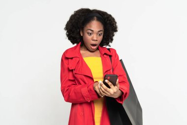 Young shocked woman using cellphone while posing with shopping bag isolated over white background