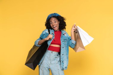 Black woman expressing surprise while posing with shopping bags isolated over yellow background