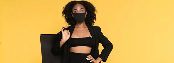 Black woman in face mask posing with shopping bag isolated over yellow background