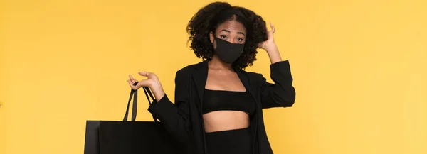 Black woman in face mask posing with shopping bag isolated over yellow background