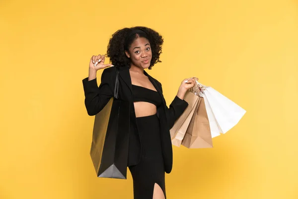 Black woman pointing fingers at herself while posing with shopping bags isolated over yellow background