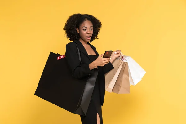 Czarny Szokujące Kobieta Pomocą Telefonu Komórkowego Podczas Pozowania Torby Zakupy — Zdjęcie stockowe