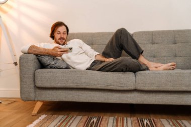 Kızıl saçlı adam evdeki kanepede dinlenirken cep telefonu kullanıyor.