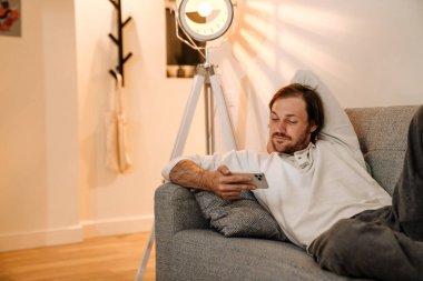 Ginger bristle man using mobile phone while resting on couch at home