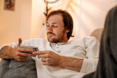 Ginger bristle man using mobile phone while resting on couch at home
