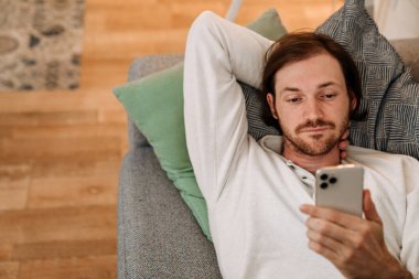 Avrupalı traşsız adam evdeki koltukta dinlenirken cep telefonu kullanıyor.