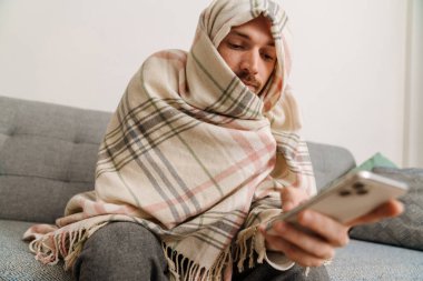 European man wrapped blanket using cellphone while sitting on sofa at home