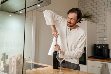 Avrupalı traşsız adam evde şarap şişesi açıyor.