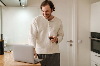 Avrupalı traşsız adam evde kırmızı şarap içerken dizüstü bilgisayar kullanıyor.