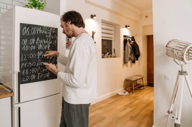 Avrupalı bir adam mutfaktaki masanın başında beklerken cep telefonu kullanıyor.