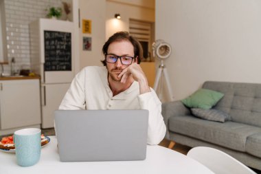 Gözlüklü kızıl saçlı adam evde yemek yerken bilgisayarla çalışıyor.