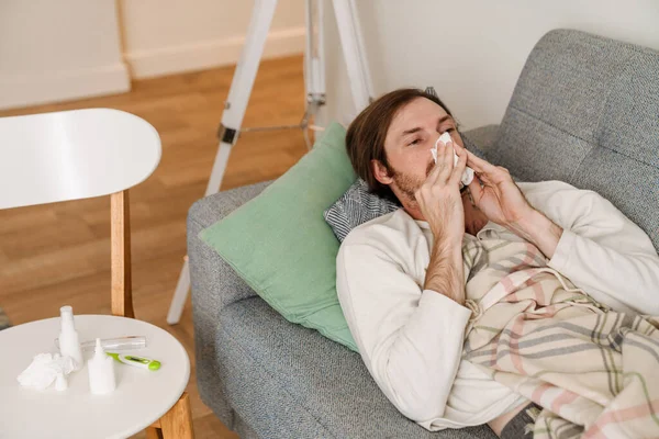 Ginger man feeling sick and sneezing while lying on couch at home