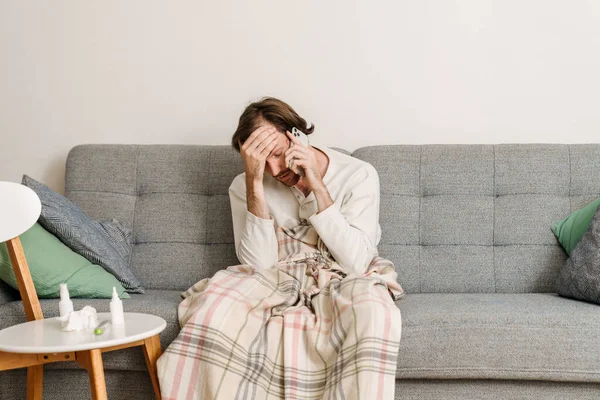 Grip olmuş Avrupalı bir adam evde koltukta otururken cep telefonuyla konuşuyor.