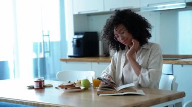 Smiling curly haired woman in eyeglasses looking at phone in the morning at home