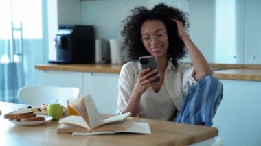 Cheerful curly haired woman in eyeglasses drinking orange juice and texting on phone in the morning at home