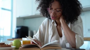 Pleased curly haired woman in eyeglasses drinking orange juice and reading book in the morning at home