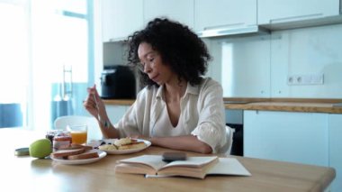 Pretty curly haired woman in eyeglasses eating breakfast in the morning at home