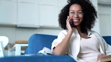 Pretty curly haired woman in eyeglasses talking on phone while sitting on the sofa at home