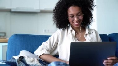 Cheerful curly haired woman in eyeglasses talking by video call on laptop while sitting on the sofa at home