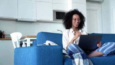 Surprised curly haired woman in eyeglasses talking by video call on laptop while sitting on the sofa at home