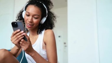 Pensive curly haired woman typing on phone in headphones at home
