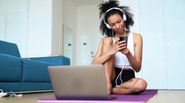 Smiling curly haired woman typing on phone in headphones on the yoga mat at home