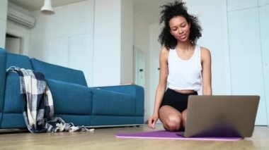 Positive curly haired woman stretching her neck and looking at laptop on the yoga mat at home