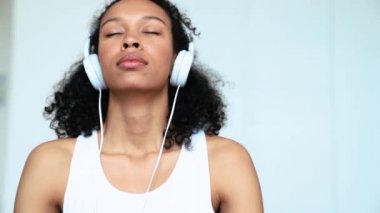 Relaxed curly haired woman doing breathing exercise and listening music in headphones on the yoga mat at home