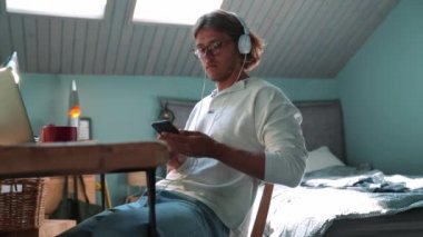 Happy blond man wearing eyeglasses listening music in headphones and typing on phone at home indoors