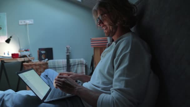 Cheerful Blond Man Wearing Eyeglasses Working Laptop While Sitting Bed — Vídeo de Stock