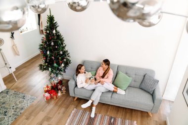 White mother and daughter smiling and talking while resting on sofa at home