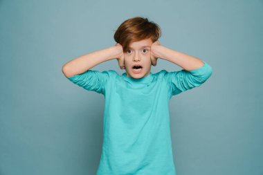 Ginger white boy expressive surprise while covering his ears isolated over blue wall