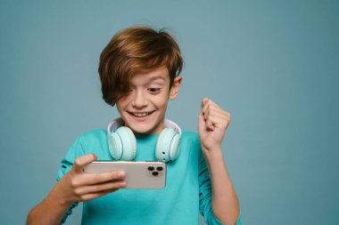 Ginger white boy gesturing while playing online game on cellphone isolated over blue wall