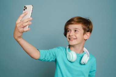 Ginger white boy with headphones taking selfie photo on cellphone isolated over blue wall