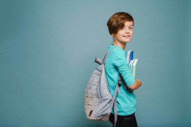 Ginger white boy smiling while posing with exercise books isolated over blue wall