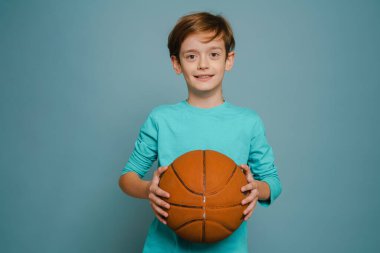 Ginger white boy smiling while posing with basketball isolated over blue wall