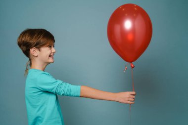 Ginger white boy smiling while posing with red balloon isolated over blue wall