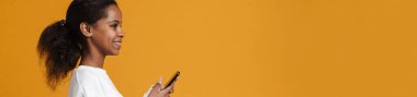 Brunette black girl smiling while using cellphone isolated over yellow background