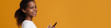 Brunette black girl smiling while using cellphone isolated over yellow background
