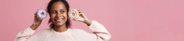 Brunette black girl laughing while posing with donuts isolated over pink background