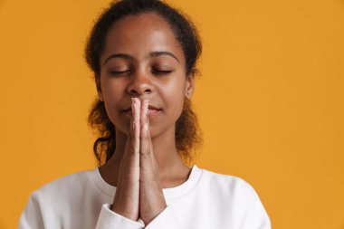 Brunette black girl meditating while holding hands together isolated over yellow background