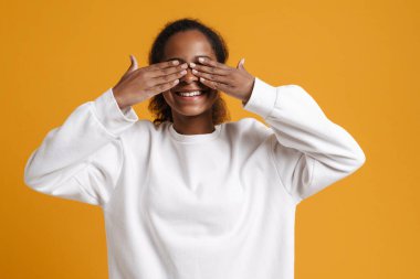Black girl wearing long-sleeve smiling while covering her eyes isolated over yellow background
