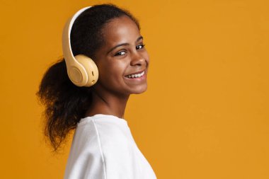 Brunette black girl smiling while listening music with headphones isolated over yellow background