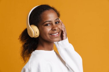 Brunette black girl smiling while listening music with headphones isolated over yellow background