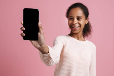 Brunette black girl smiling while showing cellphone at camera isolated over pink background