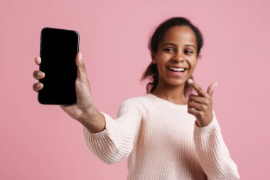 Black girl pointing finger and showing cellphone at camera isolated over pink background