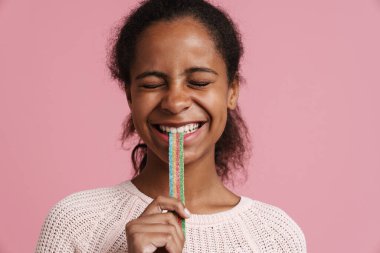 Brunette black girl smiling while eating candy worm isolated over pink background